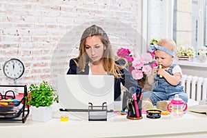 Cheerful young beautiful businesswoman looking at laptop while sitting at her working place with her little daughter