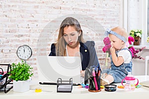 Cheerful young beautiful businesswoman looking at laptop while sitting at her working place with her little daughter