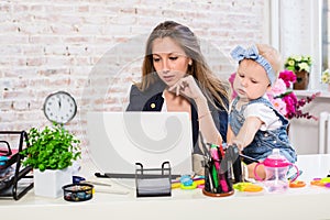 Cheerful young beautiful businesswoman looking at laptop while sitting at her working place with her little daughter