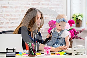 Cheerful young beautiful businesswoman looking at telephone while sitting at her working place with her little daughter