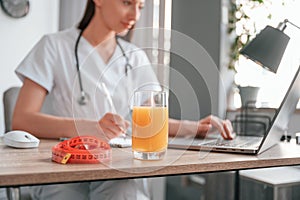 Working by using laptop. Apple juice and measuring length is on the table. Young beautiful female doctor