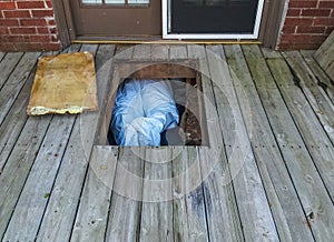 Workman with protective suit crawing under house from crawlspace underneath a wooden deck - only his legs and feet showing