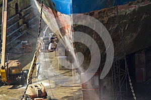 Workmen cleaning corrosion from the hull of a merchant ship with pressure sandblasting in a dry dock