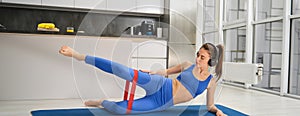 Workout and fitness concept. Woman in headphones does exercises at home, stretching rubber resistance band with legs