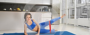 Workout and fitness concept. Woman in headphones does exercises at home, stretching rubber resistance band with legs