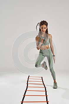 Workout. Full length shot of young sporty mixed race woman in sportswear training on agility ladder drill isolated over