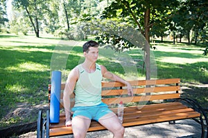 Workout is over. Man with yoga mat and water bottle sit on bench in park. Join outdoors yoga practice. Athlete with yoga