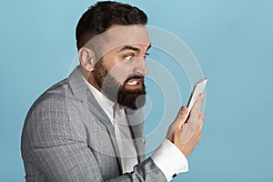 Workplace anger management. Pissed young businessman looking at smartphone screen on blue background