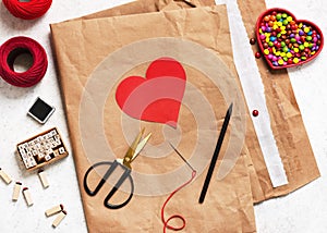 Workplace with crafts equipment: used shipping bag, red heart template, pen, scissors, letter stamps, colorful sweet, needle