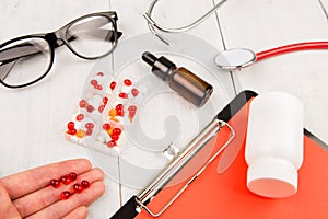 Workplace of doctor. Pills in hand, red stethoscope, clipboard and glasses on white wooden table