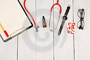 Workplace of doctor. Red stethoscope, clipboard, glasses, watch and pills on white wooden table