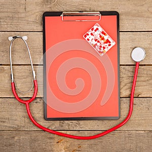Workplace of a doctor. Red stethoscope, clipboard and pills on wooden desk