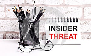 A workplace in the house with glasses, pencils in a stand and a notebook with text INSIDER THREAT on a brick wall background. Home
