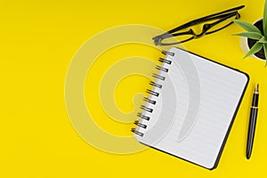 Workspace with black pen, spectacles, decoration flower and book on yellow background