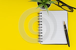 Workspace with black pen, spectacles, decoration flower and book on yellow background