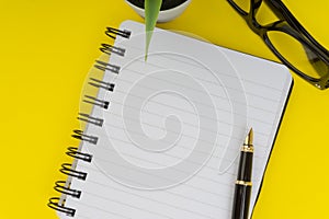 Workspace with black pen, spectacles, decoration flower and book on yellow background