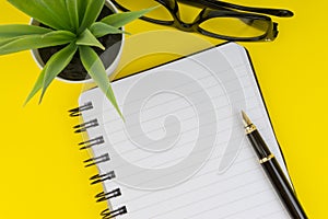 Workspace with black pen, spectacles, decoration flower and book on yellow background