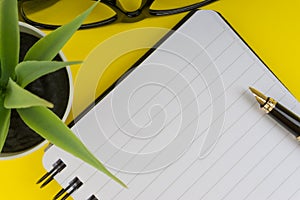 Workspace with black pen, spectacles, decoration flower and book on yellow background