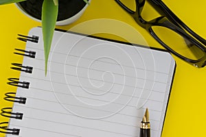 Workspace with black pen, spectacles, decoration flower and book on yellow background