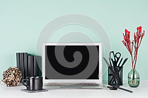 Workspace with blank laptop monitor, black stationery, books, round sheaf of dry brown branches, coffee cup, red plant in  green