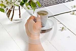 Workspace with girl`s hand on laptop keyboard and cup of coffee, white spring apple tree flowers on white woodden background. Vie