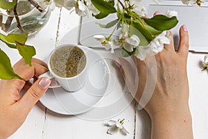 Workspace with girl`s hand on laptop keyboard and cup of coffee, white spring apple tree flowers on white woodden background. Vie