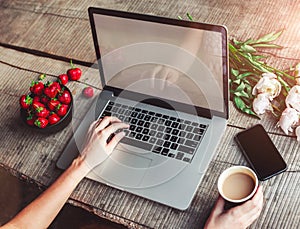 Workspace with girl`s hands, laptop computer, bouquet of peonies flowers, coffee, strawberries