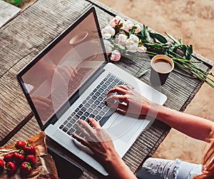 Workspace with girl`s hands, laptop computer, bouquet of peonies flowers, coffee, strawberries