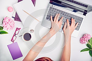 Workspace with girl`s hands on laptop keyboard, notebook, glasses, cup of coffee and wisteria flowers on white background. Top vi