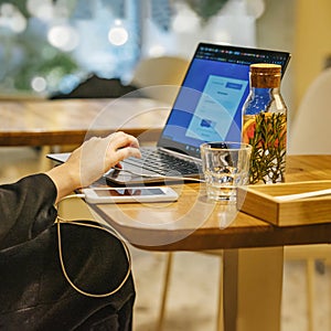 Workspace inside cafe, girl hands, laptop computer, mobile phone, vitamin drink on table. Lifestyle, modern technology