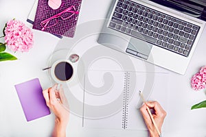 Workspace with laptop, girl`s hands writing in notebook, glasses, cup of coffee and wisteria flowers on white background. Top vie