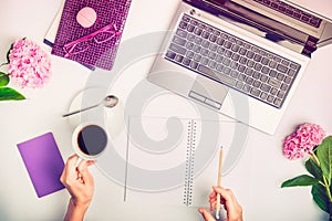 Workspace with laptop, girl`s hands writing in notebook, glasses, cup of coffee and wisteria flowers on white background. Top vie photo