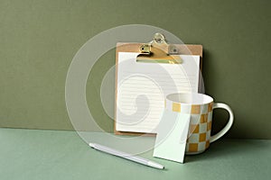 Workspace notepad binder clip, pen, mug cup on green desk. khaki wall background