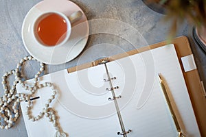 Opened notebook with clean sheets, gold pen, rosary, pink colored case and cup of tea near vase