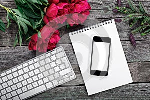 Workspace with smartphone, keyboard, notepad, fir branch and peonies flowers bouquet on rustic background