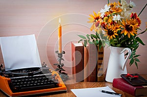 Workspace with vintage orange typewriter. Bouquet of summer flowers, candles and old books