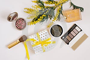 A workspace with yellow mimosa flowers, decorative cosmetics, a gift box with a bow on a white background.