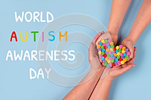 World Autism Awareness Day concept - autistic child`s hands supported by mother holding multicolored heart on blue background.