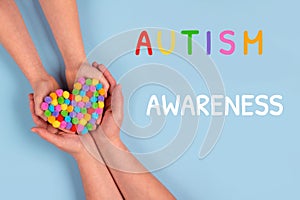 World Autism Awareness Day concept - autistic child`s hands supported by mother holding multicolored heart on blue background.
