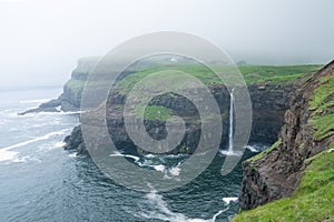 World famous Mulafossur waterfall falling from the cliff near Gasadalur village into the ocean in Faroe Islands . Misty, cloudy