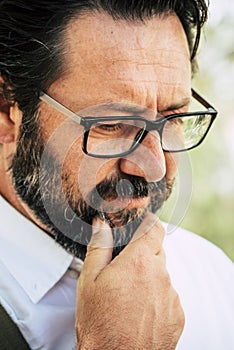 Worried adult caucasian 50 years old man touch the beard and think alone - eyeglasses and people outdoor with green park defocused