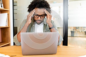 Worried african-american male employee colleague staring at laptop screen sitting on the workplace