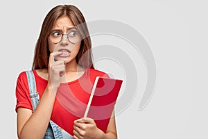 Worried anxious young woman keeps index finger near mouth, looks desperately aside wears round spectacles, carries red