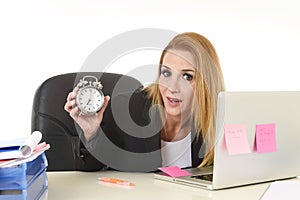 Worried attractive blond businesswoman holding alarm clock sitting at office desk working on laptop