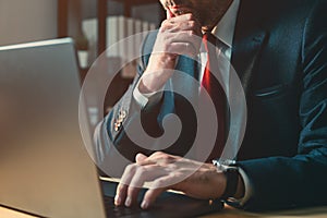 Worried businessman working on laptop computer late at night in dark office, thinking with hand on the chin