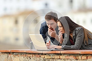 Worried couple checking news on a laptop in a terrace