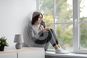 Worried depressed anxiety millennial female sitting on windowsill at home and talking on phone, calling to boyfriend