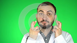 Worried doctor man standing with fingers crossed for good luck. Green screen background