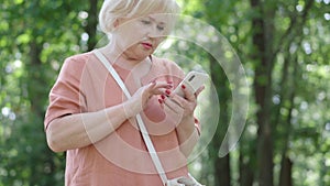 Worried embarrassed senior woman looking around in sunny park or forest and checking online map. Portrait of lost