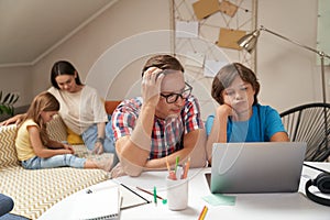 Worried father helping his little son with homework while mother and daughter sitting on sofa and playing with pazzles